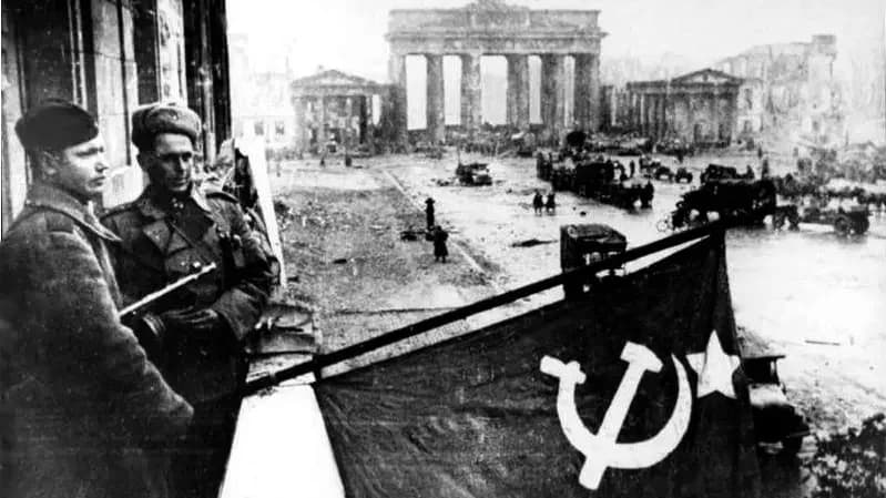 Soldados russos segurando a bandeira soviética com o portão de Brandemburgo ao fundo, no centro de Berlim.