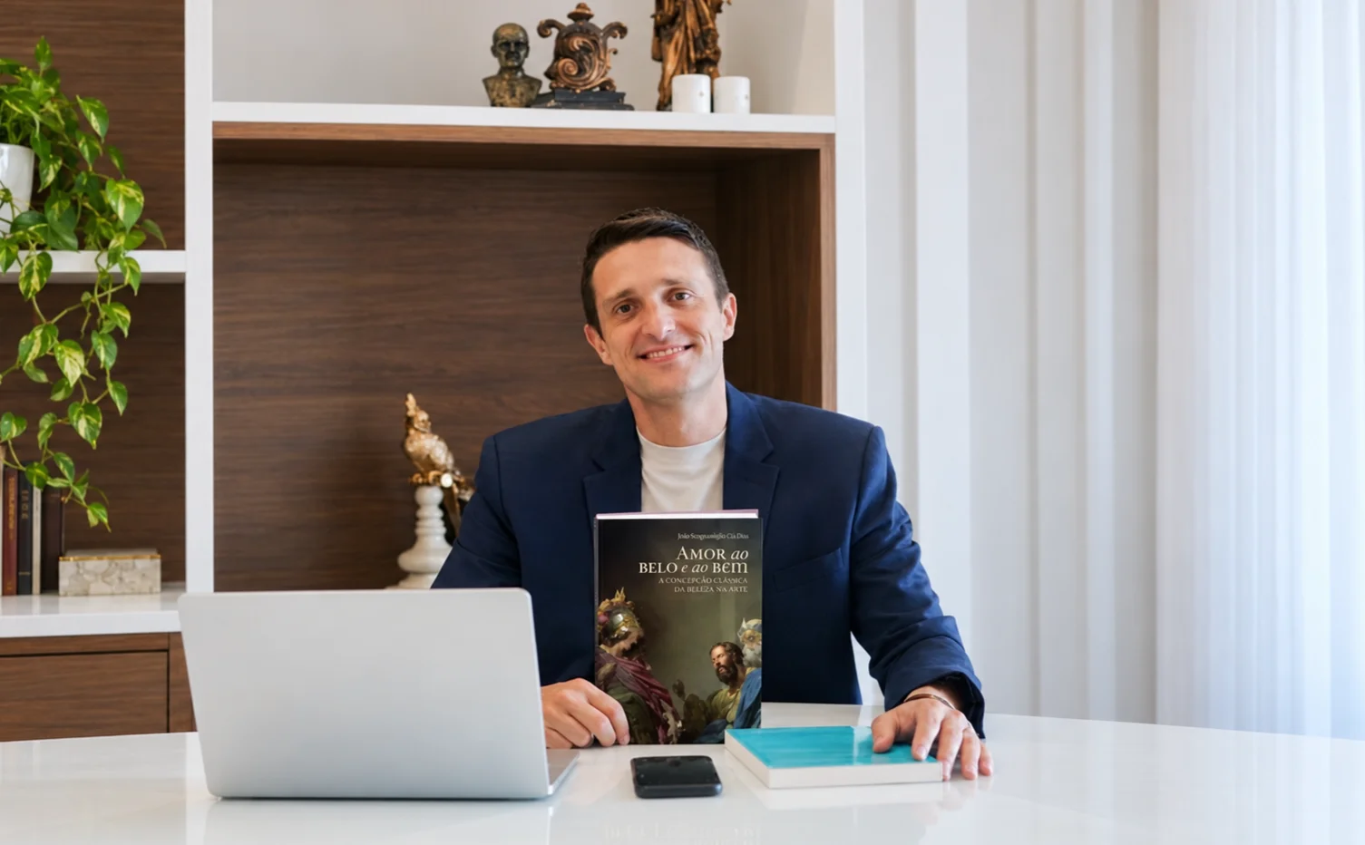 Homem sorridente sentado à mesa segurando um livro em um escritório
