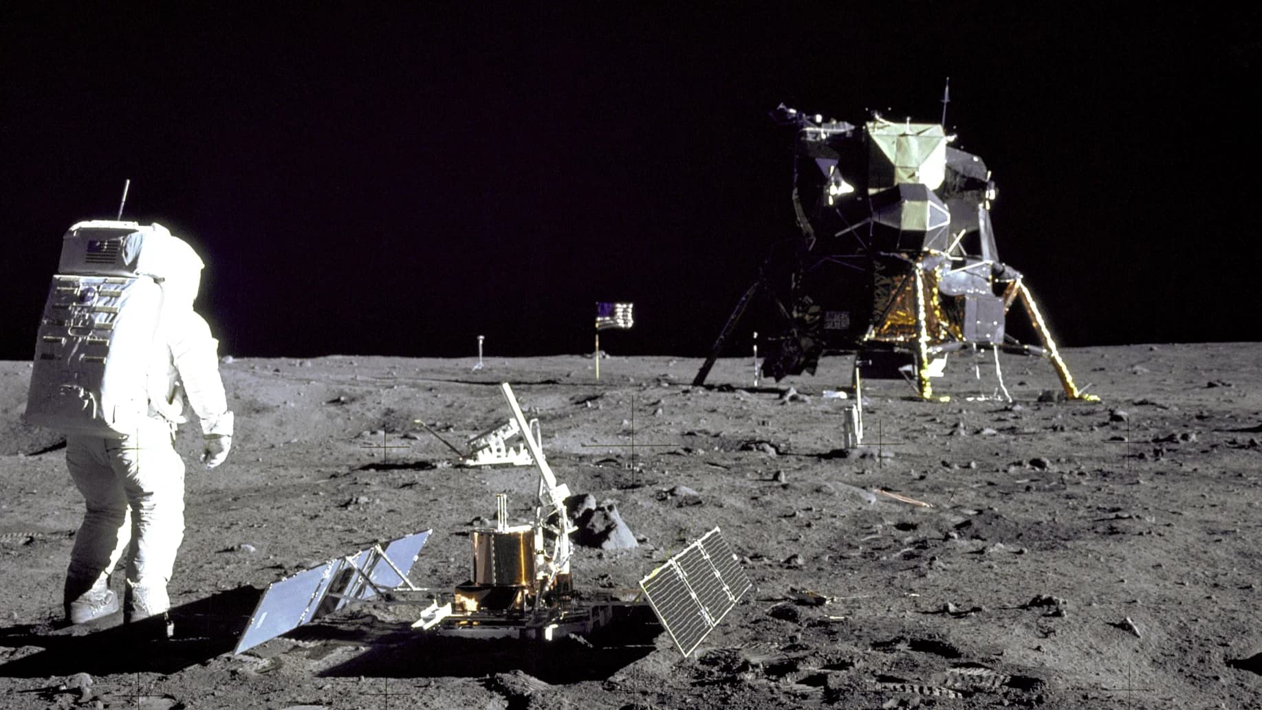 O astronauta Buzz Aldrin na superfície da Lua durante a missão Apollo 11. Ao centro, equipamentos científicos; ao fundo, o Módulo Lunar Eagle e a bandeira dos EUA sob um céu negro.