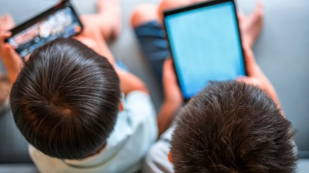 Duas crianças usam celular e tablet sentadas no sofá