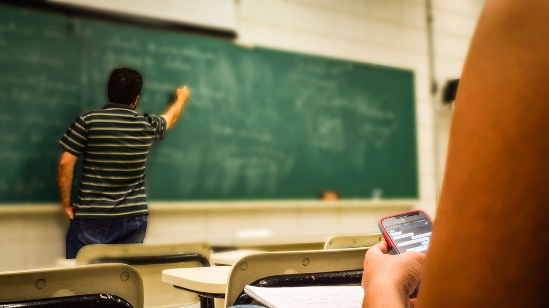 Aluna usando celular em sala de aula durante explicação do professor.