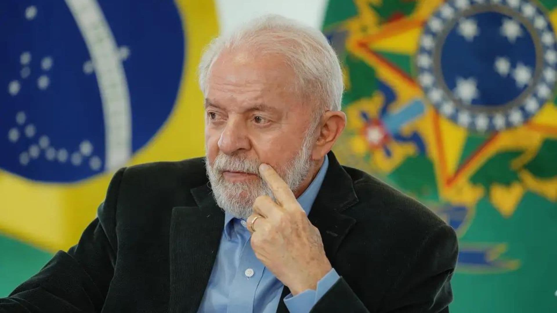 Presidente Lula mexendo na barba em frente a bandeira do Brasil e brasão de armas da República