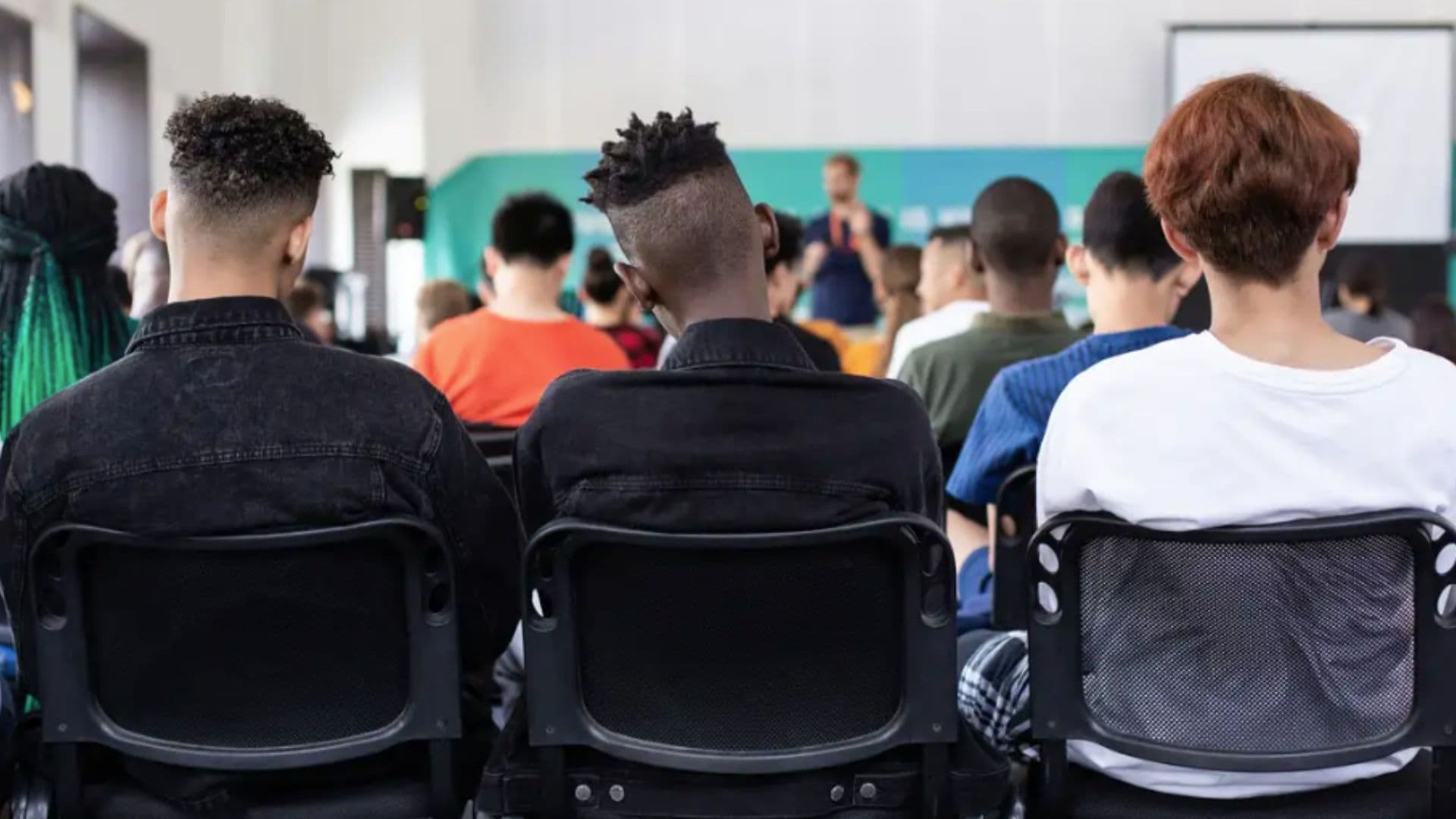 Jovens aparecem de costas, enquanto assistem aula