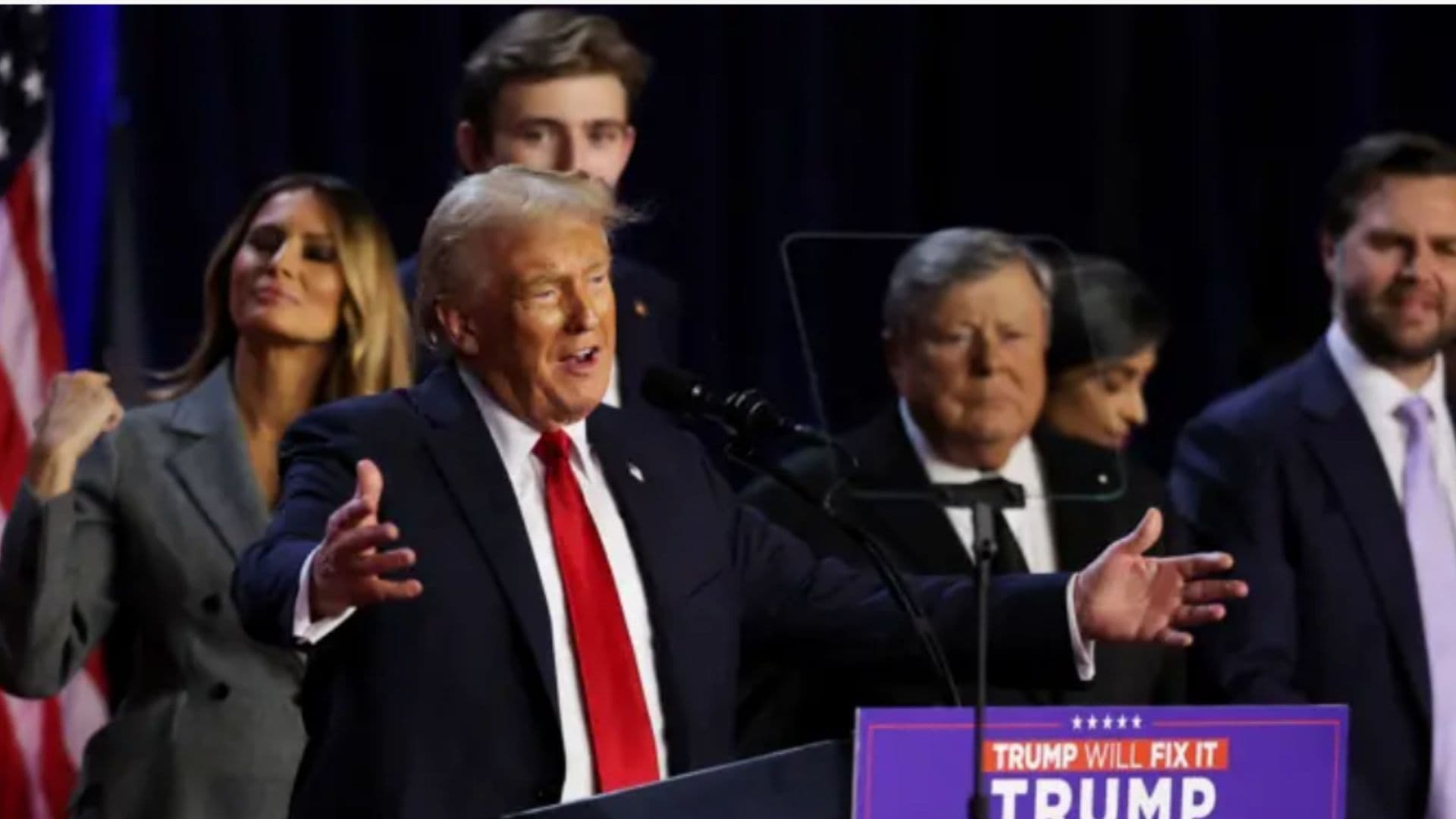 Donald Trump aparece em primeiro plano comemorando sua vitória eleitoral. Atrás a sua direita está sua esposa Melania Trump e seu filho Barron Trump. Trump promete liberar réus do 6 de janeiro