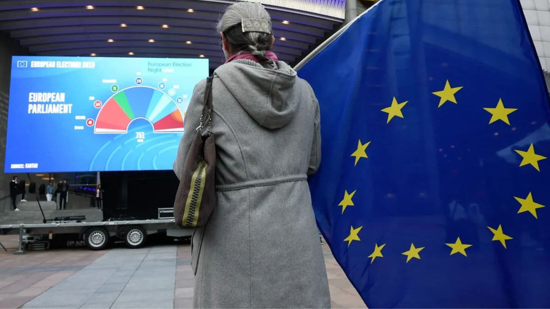 Mulher segurando a bandeira da União Europeia em frente a um gráfico com a composição do Parlamento Europeu
