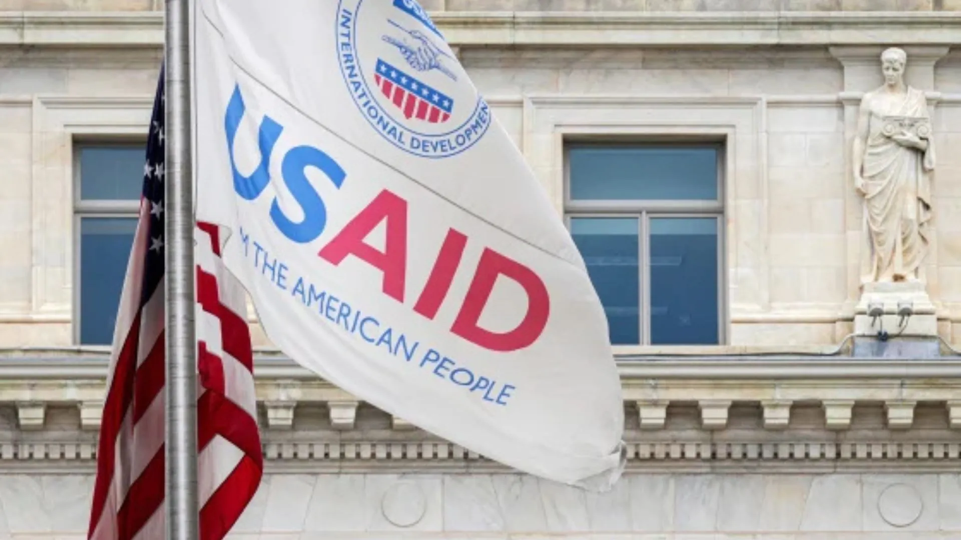 Fachada do prédio onde ficava o escritório da USAID com foco em uma bandeira da agência hasteada no mesmo mastro que uma dos EUA