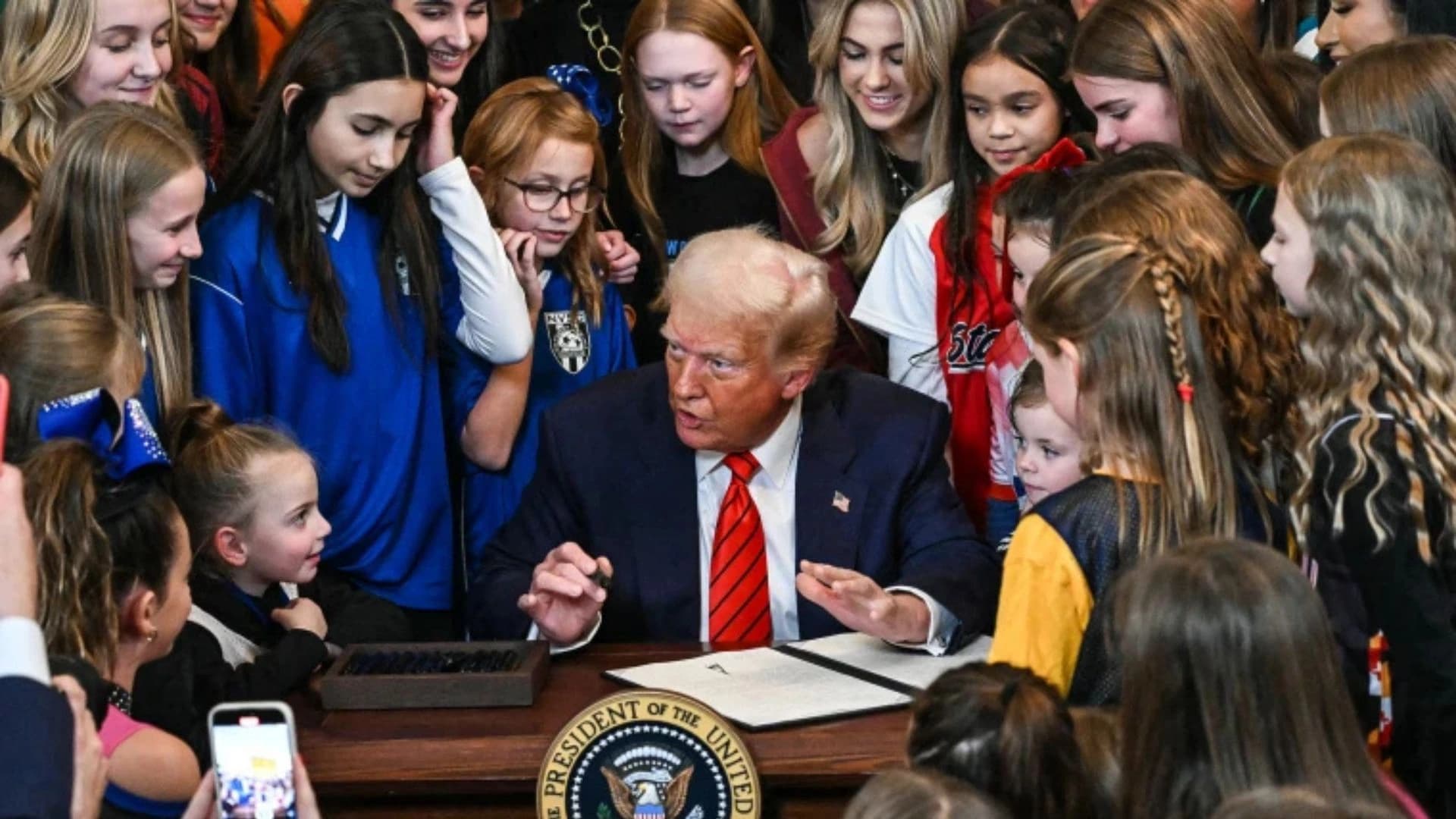 Trump assinando decreto sobre atletas trans cercado de garotas com roupas de esporte.